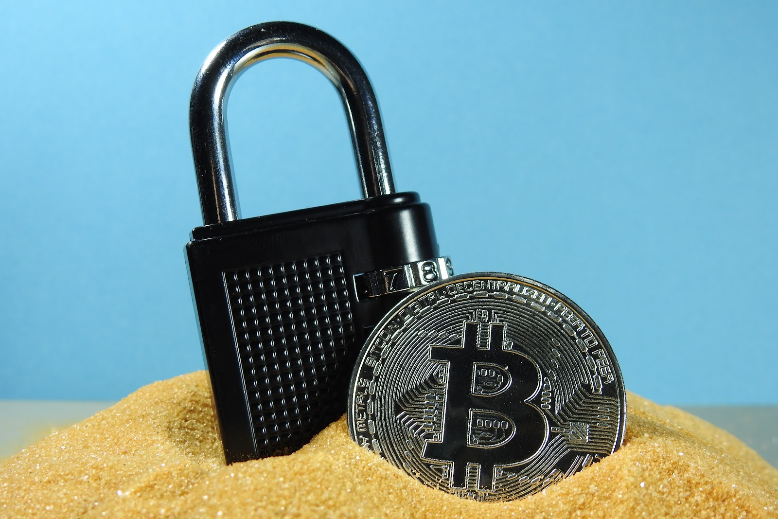 a padlock and a bitcoin on sand in front  of a blue background